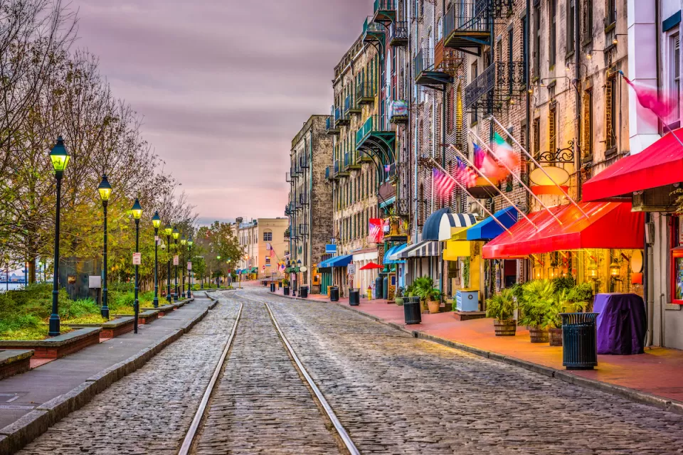 Cobblestone street with streetcar line, row of vintage streetlights on left, and colorful restaurant and shop awnings on right on River Street in Savannah, Georgia.